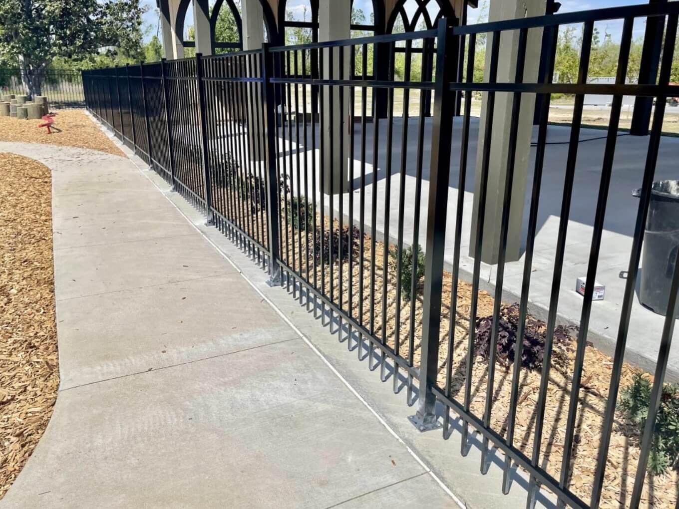 Black aluminum picket fence along a concrete sidewalk beside a modern building with arched openings