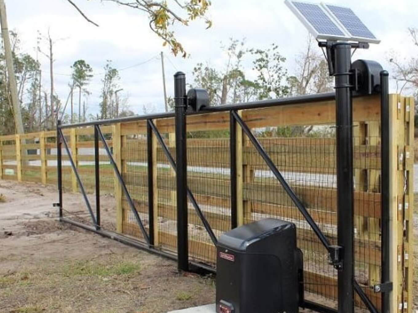 Motorized driveway gate with black frame and wire mesh set in a wood fence, with a solar panel on a post