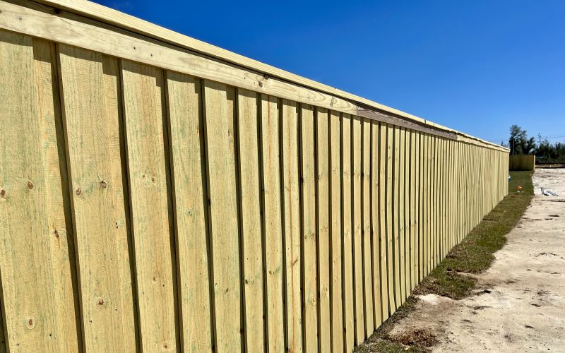 Long, newly installed vertical wood privacy fence on sandy ground under a clear sky