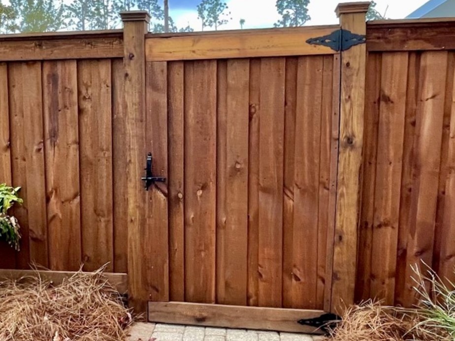 Medium-brown wood privacy fence and gate with black strap hinge and thumb latch beside a brick paver walkway