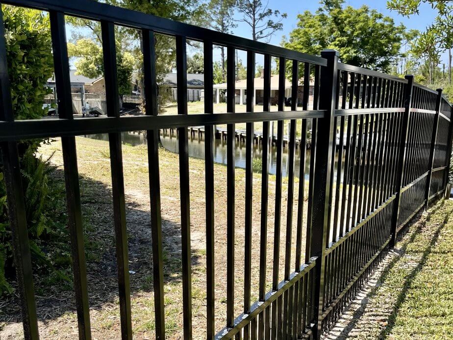 Black ornamental aluminum fence along a grassy edge with a body of water and homes beyond