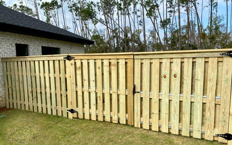 Light wood shadowbox-style privacy fence with a wide double gate and heavy black hinges on a manicured lawn
