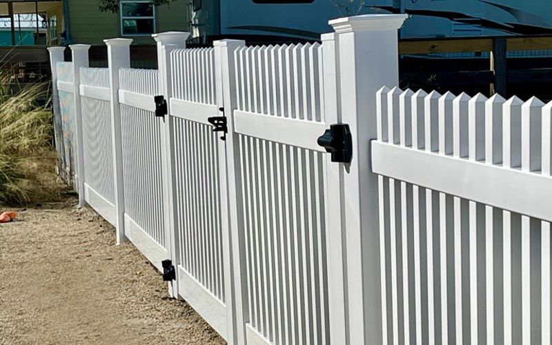 White vinyl semi-privacy fence with pyramid post caps and black gate hardware on light sandy ground