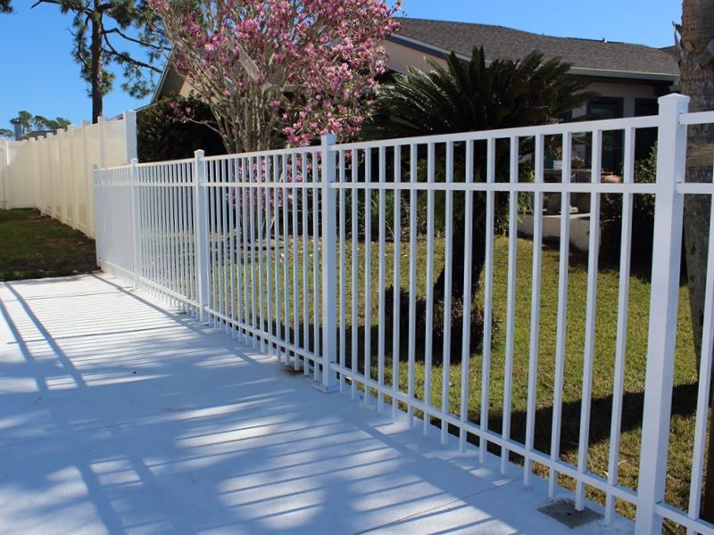 White aluminum picket fence along a white patio with palm trees and shrubs